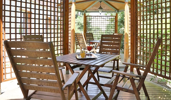 Una terraza de madera con una mesa y sillas, un vaso de bebida roja y una bandeja con aperitivos.
