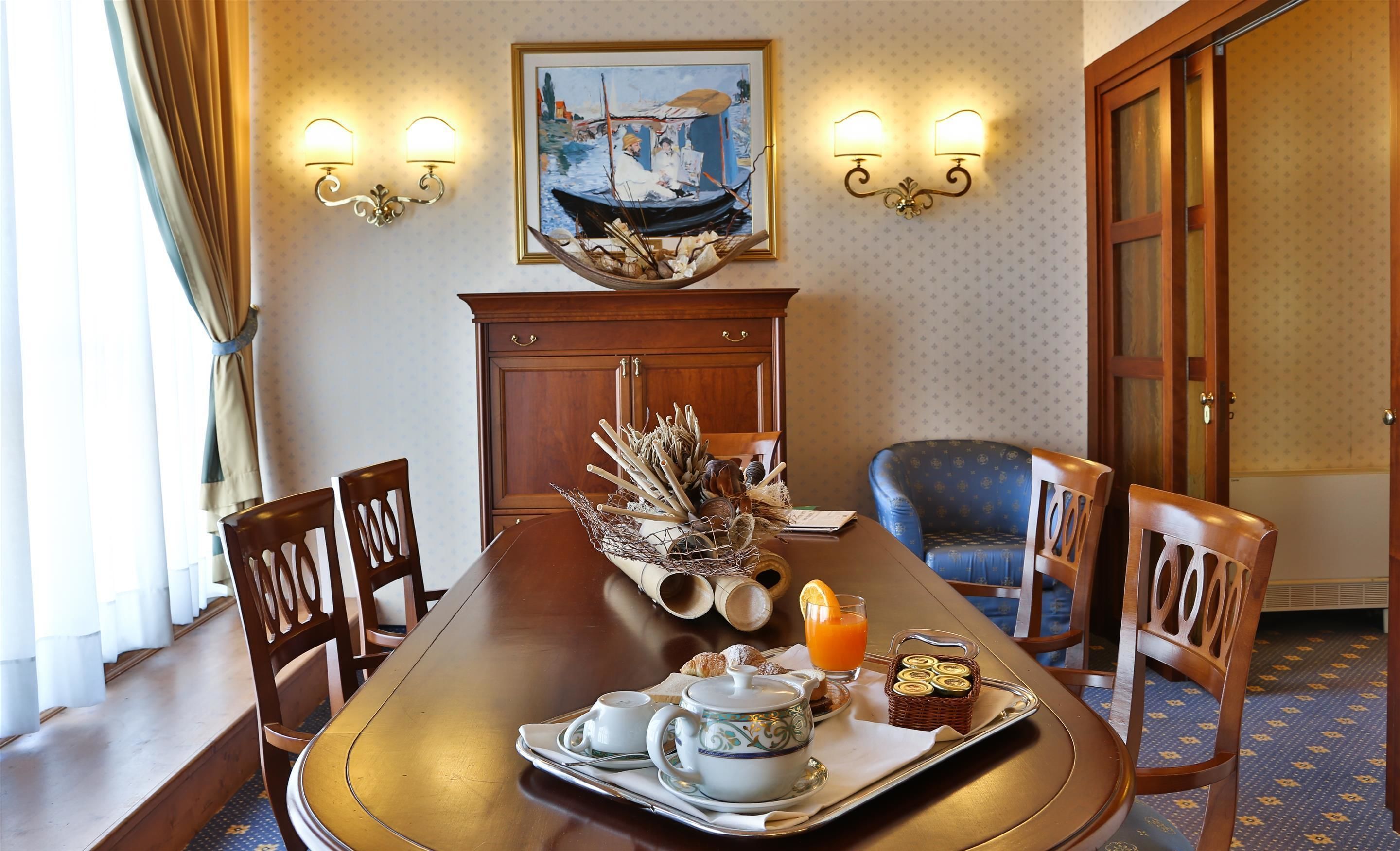 Un comedor con una mesa de madera dispuesta para el desayuno, que incluye una tetera, tazas y pasteles. Hay una silla con un cojín azul y una cesta decorativa sobre la mesa.