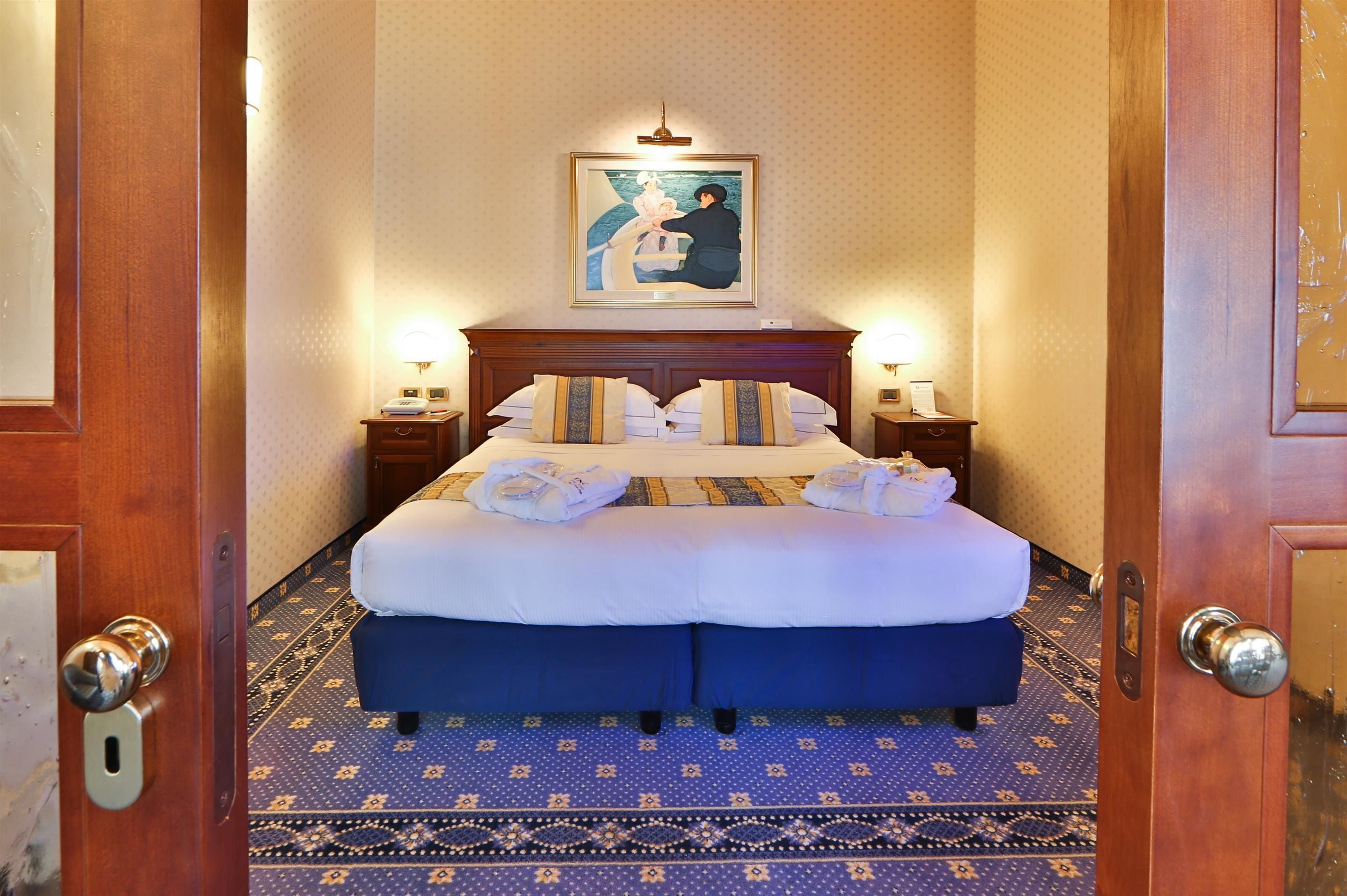 A hotel room with a large bed, two bedside tables, a painting on the wall, and a patterned carpet.