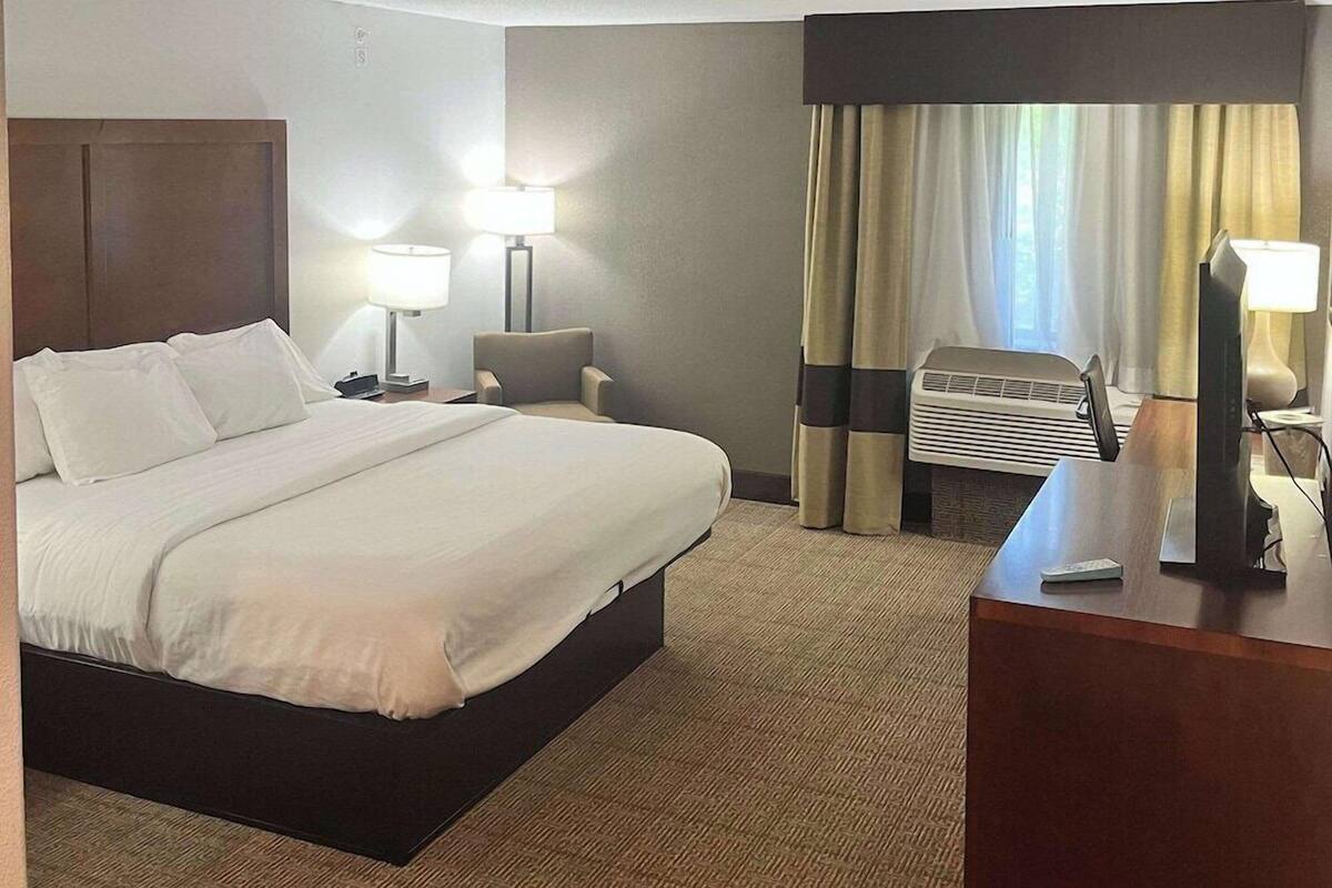 A hotel room with a bed, two bedside lamps, a desk, a chair, a window with curtains, and an air conditioning unit.