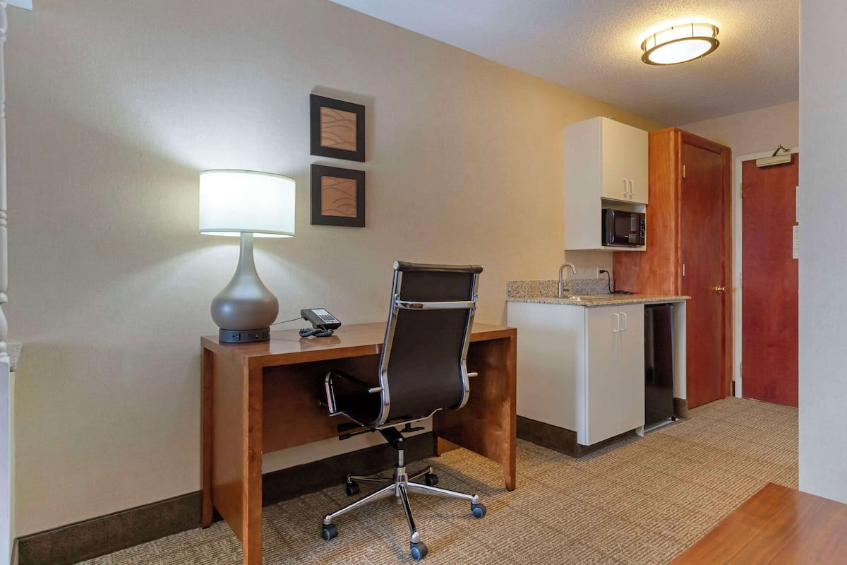 A hotel room with a wooden desk, a lamp, a chair, a microwave, and a small kitchenette.