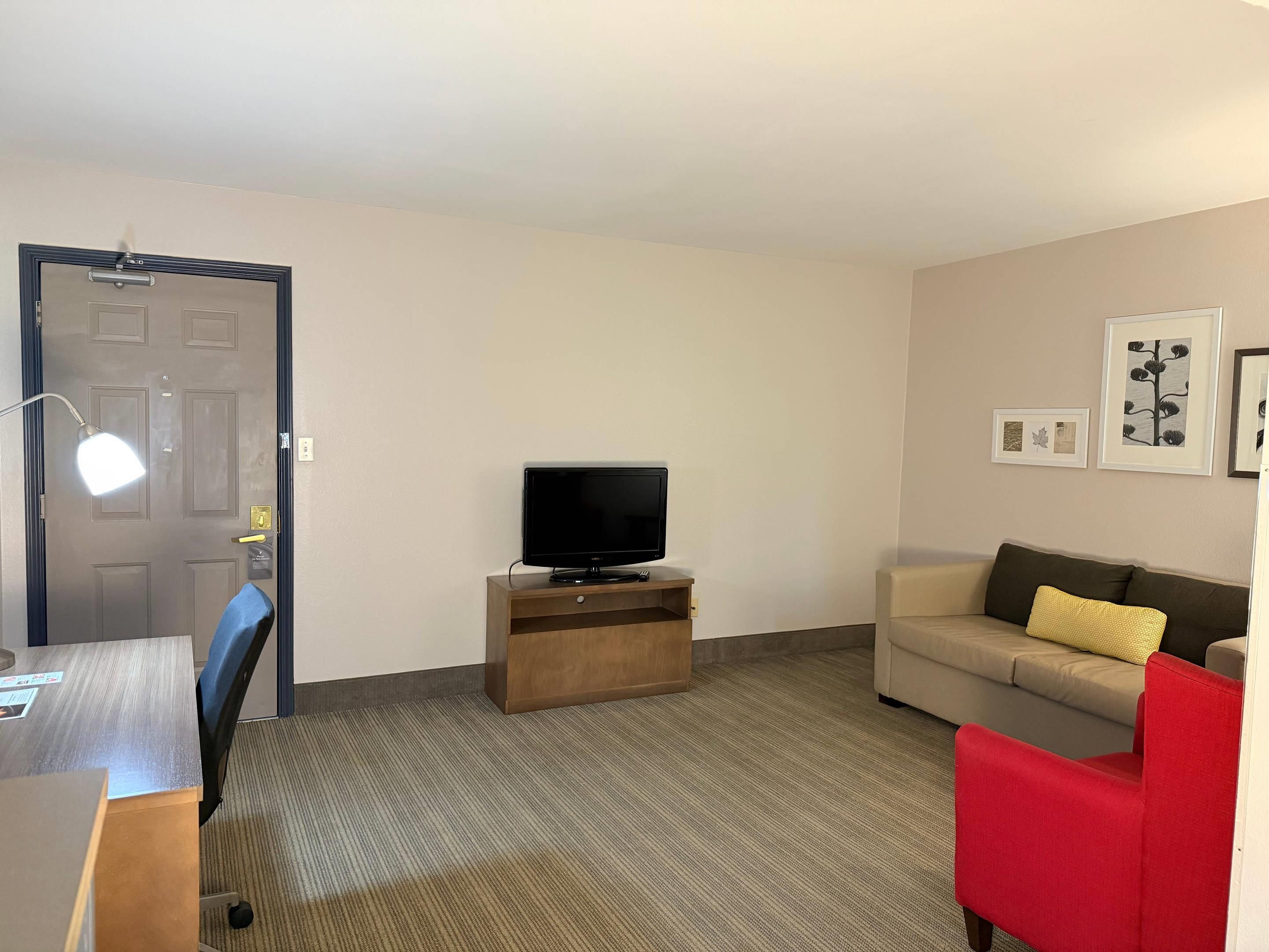 A hotel room with a flat-screen TV on a wooden stand, a beige sofa with a yellow pillow, a red chair, a desk with a chair, and a door with a handle and a light fixture.