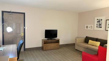 A hotel room with a flat-screen TV on a wooden stand, a beige sofa with a yellow pillow, a red chair, a desk with a chair, and a door with a handle and a light fixture.