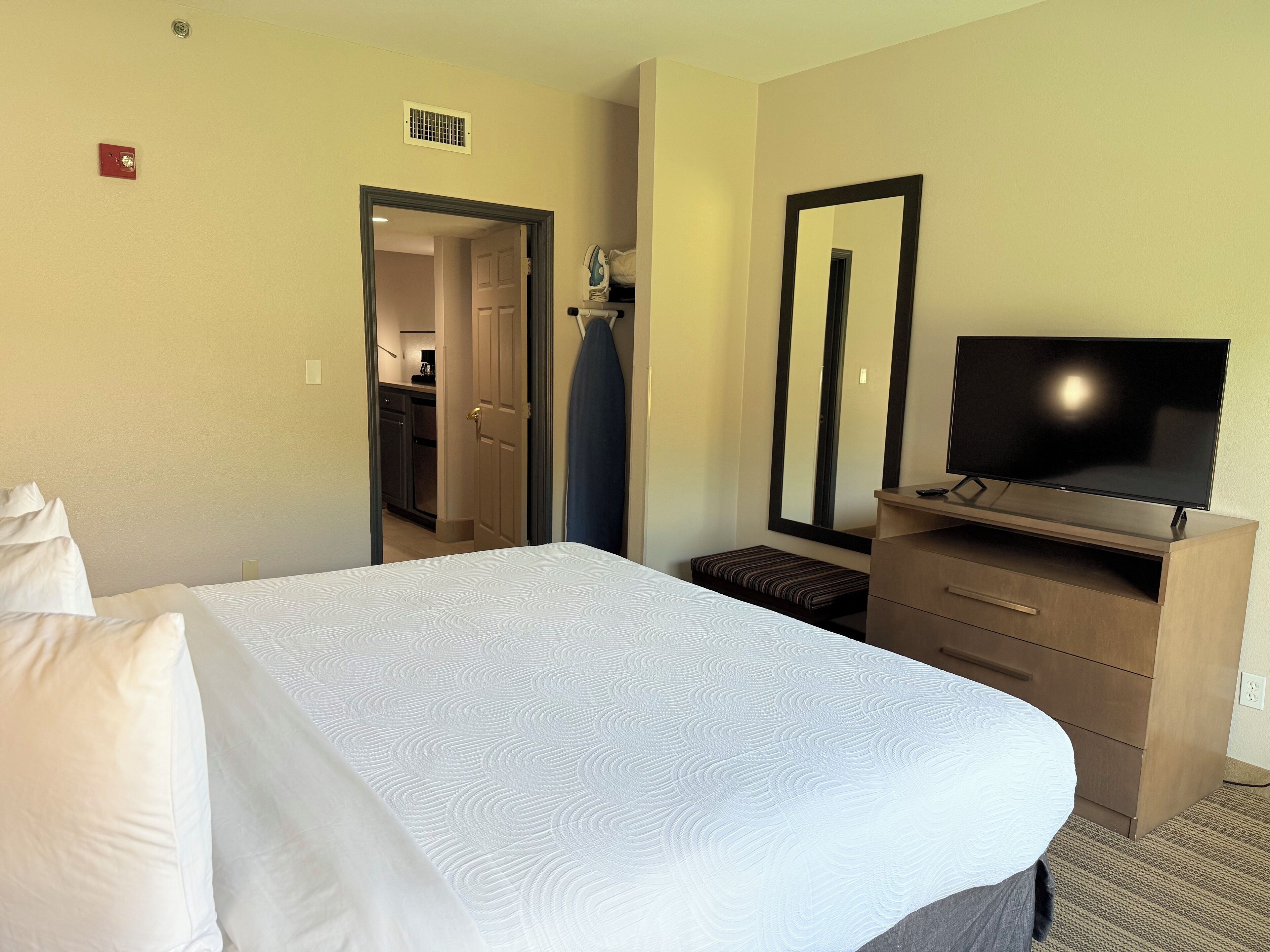 A hotel room with a large bed, a TV on a wooden dresser, a mirror, and a door leading to a kitchenette.