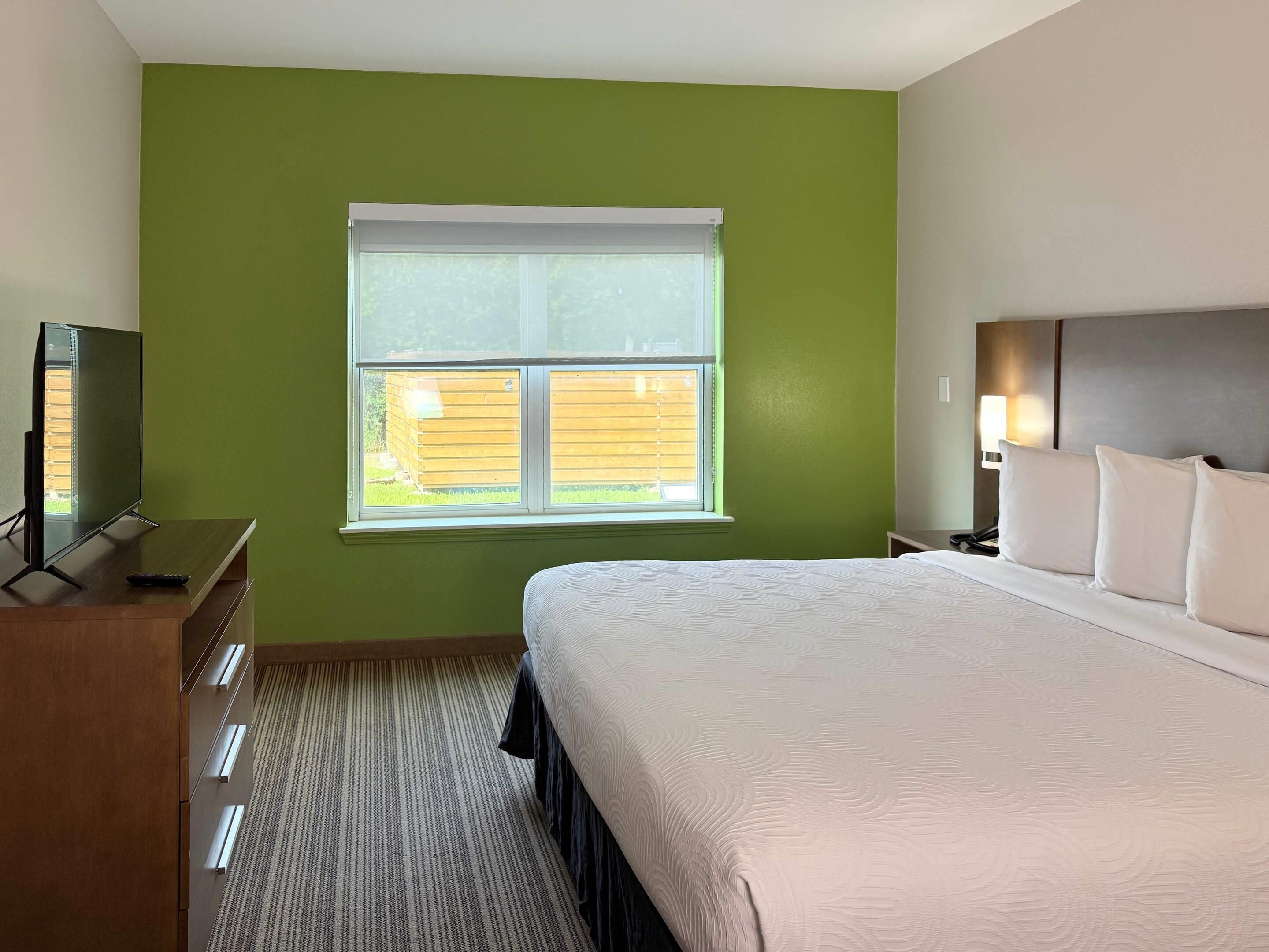 Una habitación de hotel con una cama grande, un televisor sobre un tocador de madera, una ventana con persianas y una mesita de noche con una lámpara.