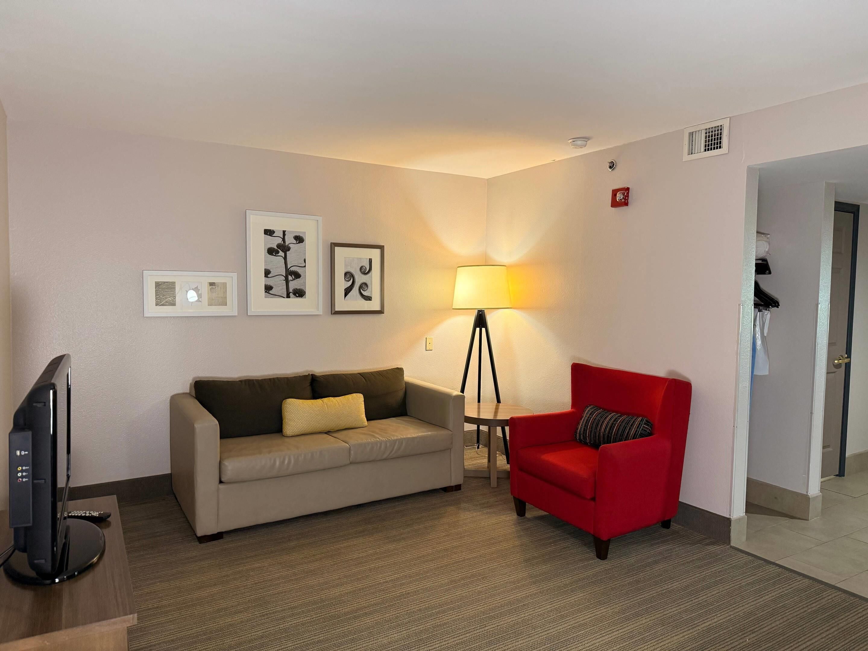 A hotel room with a beige sofa, a red armchair, a floor lamp, and a flat-screen TV on a wooden stand.