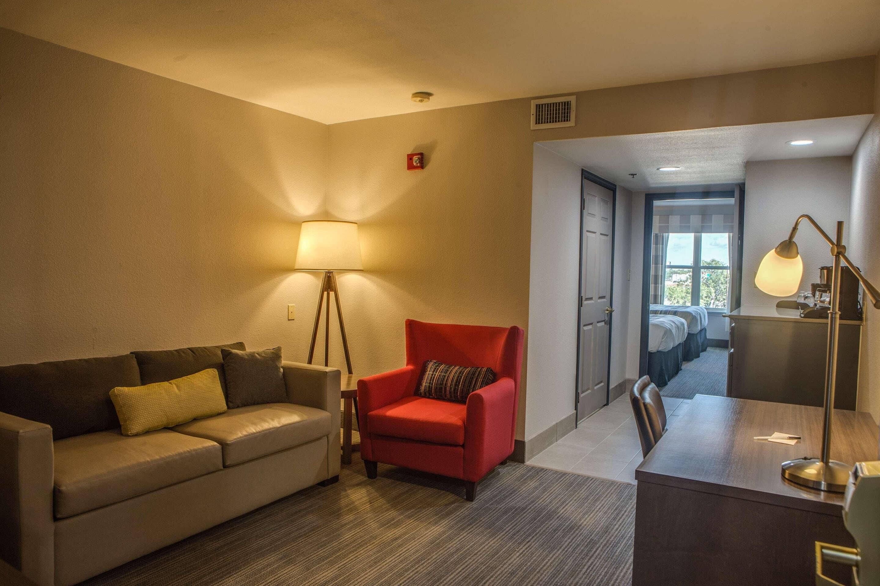 A hotel room with a sofa, armchair, desk, lamp, and a view of the corridor and another room.
