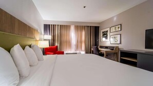 Ein Hotelzimmer mit einem großen Bett, einem Schreibtisch, einem roten Sessel, einem Fernseher und Vorhängen.