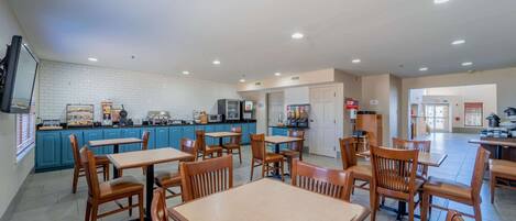 A spacious dining area with multiple wooden tables and chairs, a kitchen area with blue cabinets, and a clean, well-lit environment.
