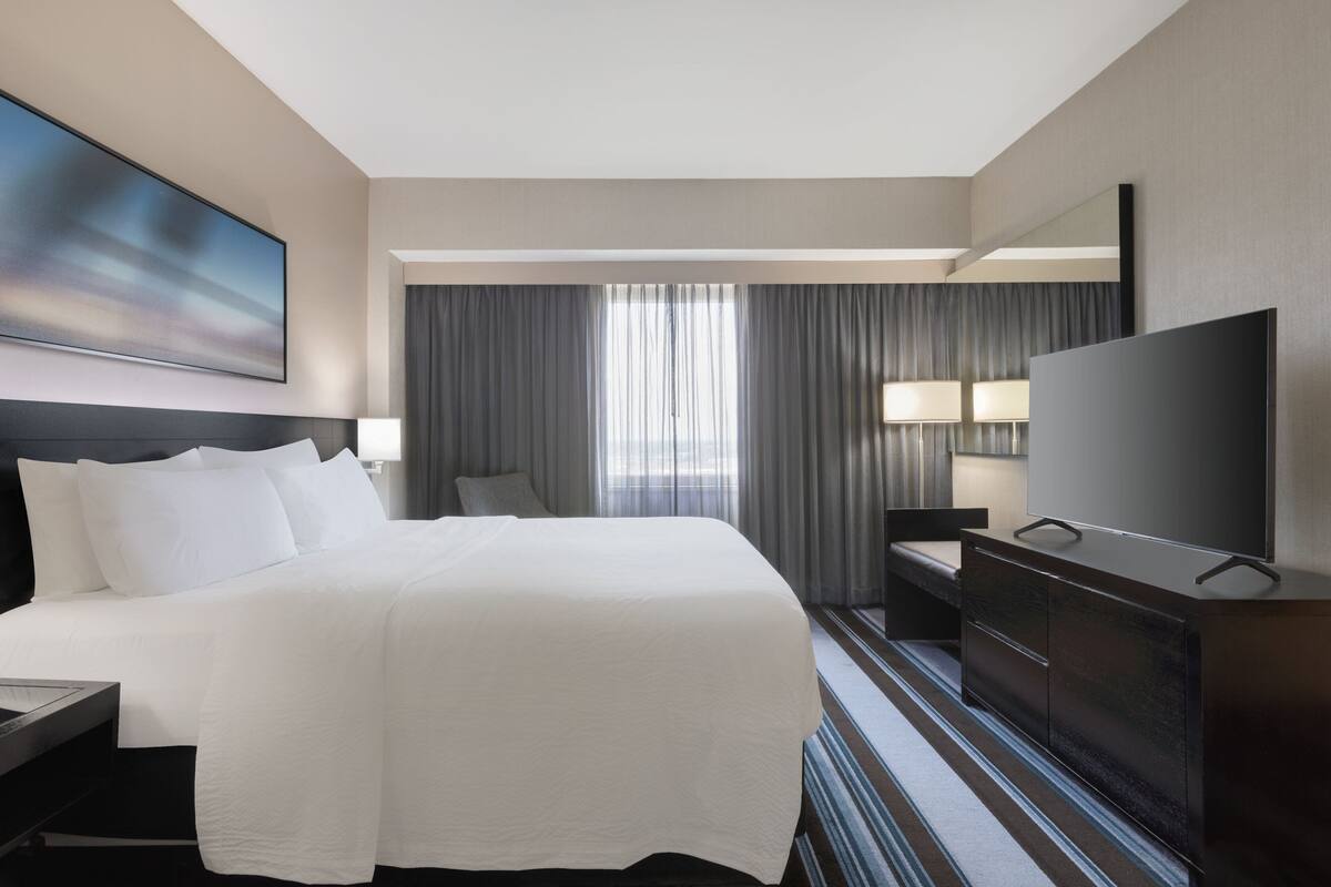 A hotel room with a large bed, a flat-screen TV, a desk with a lamp, and a large window with curtains.