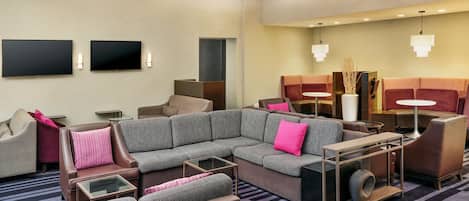 A modern hotel lobby with a sectional sofa, armchairs, a small table, and a flat-screen TV mounted on the wall.