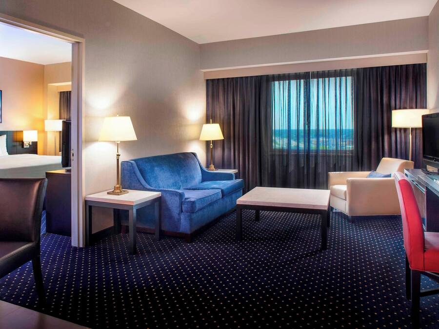 Habitación de hotel con sofá azul, una mesita, una silla roja, televisión y vista al océano a través de amplias ventanas.