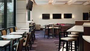 A modern hotel lobby with round tables and bar stools, a sofa area, and a large TV screen. - Courtyard by Marriott New York JFK Airport (Jamaica)