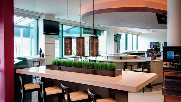 A modern hotel lobby with a central island bar, seating area, and a television displaying 'Boarding Pass'.