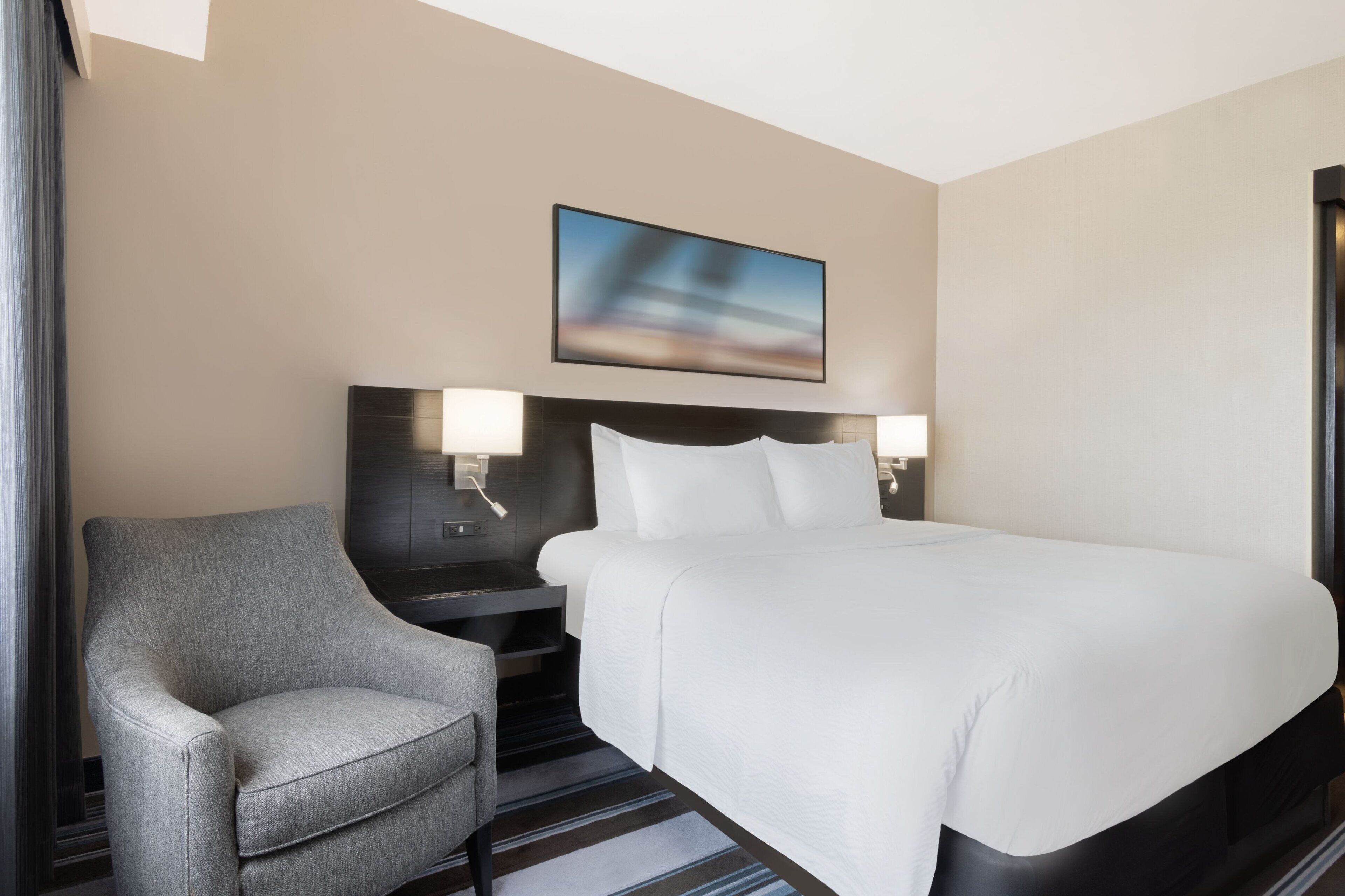 Una habitación de hotel con una cama grande, una silla, una mesita de noche y un cuadro en la pared.