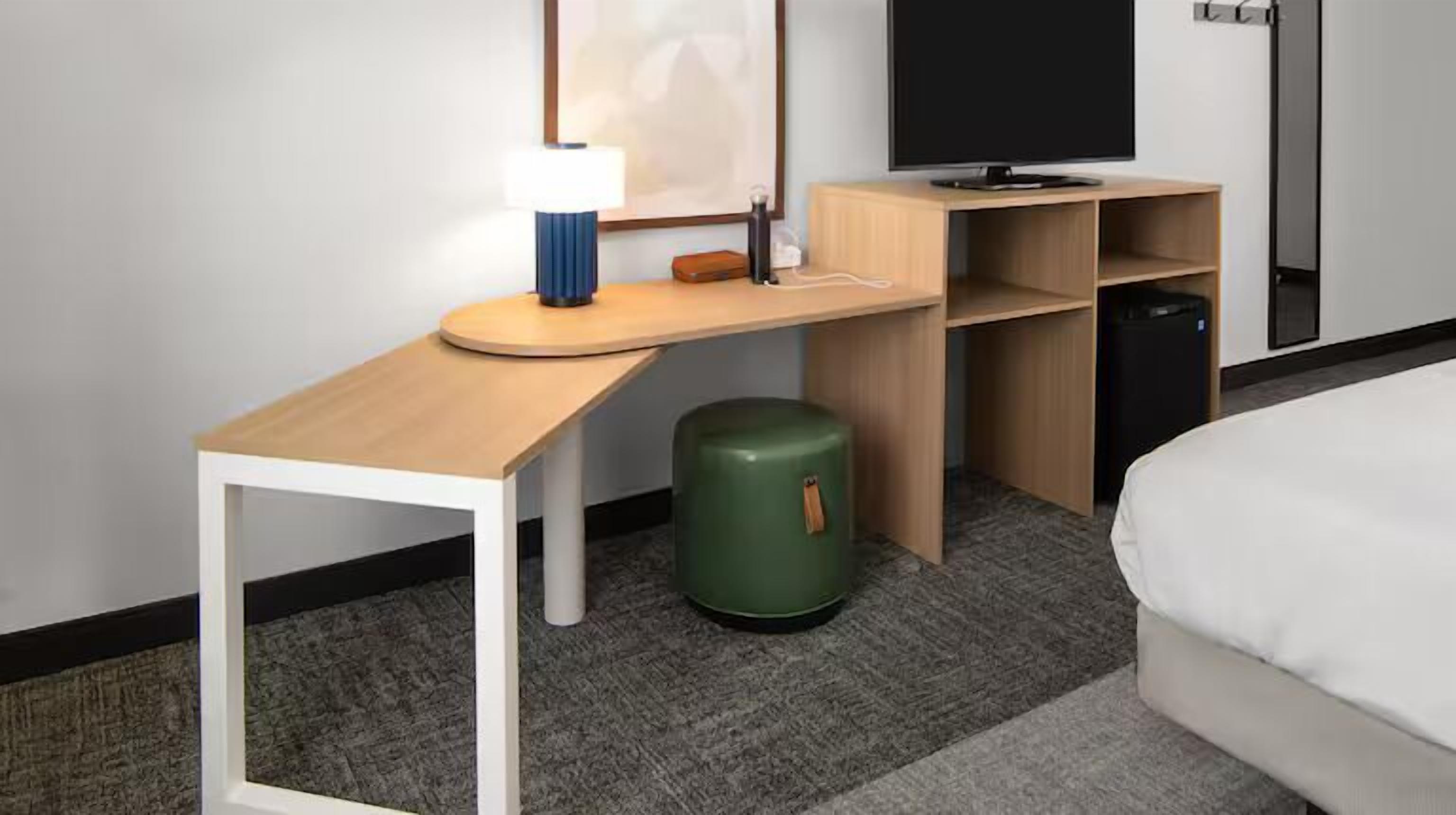 Habitación de hotel con un escritorio de madera, un bote de basura verde, una cama y un televisor sobre un mueble.