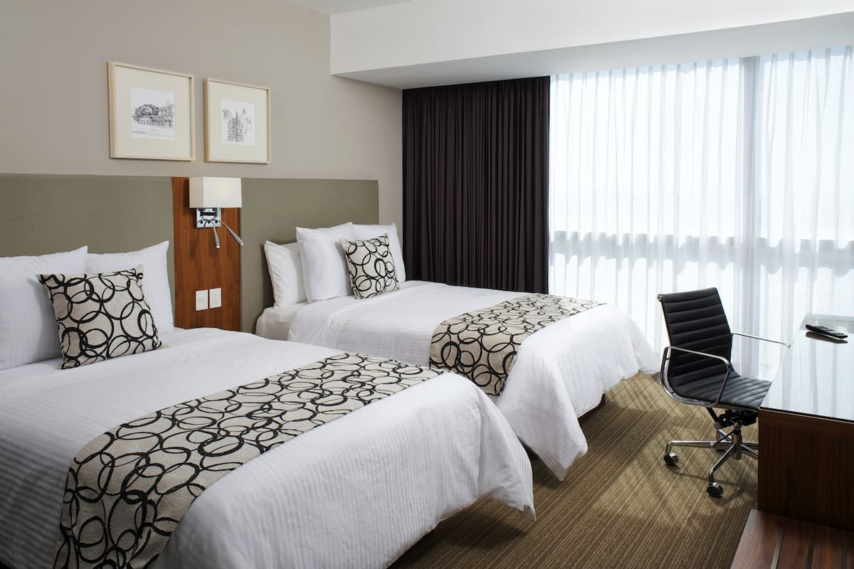 A hotel room with two beds, a desk, and a chair. There are two framed pictures on the wall.