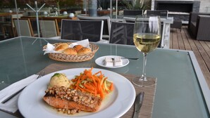A dining table with a plate of food, a glass of white wine, and a basket of bread rolls. - Casa Inn Premium Hotel Queretaro (El Marques)