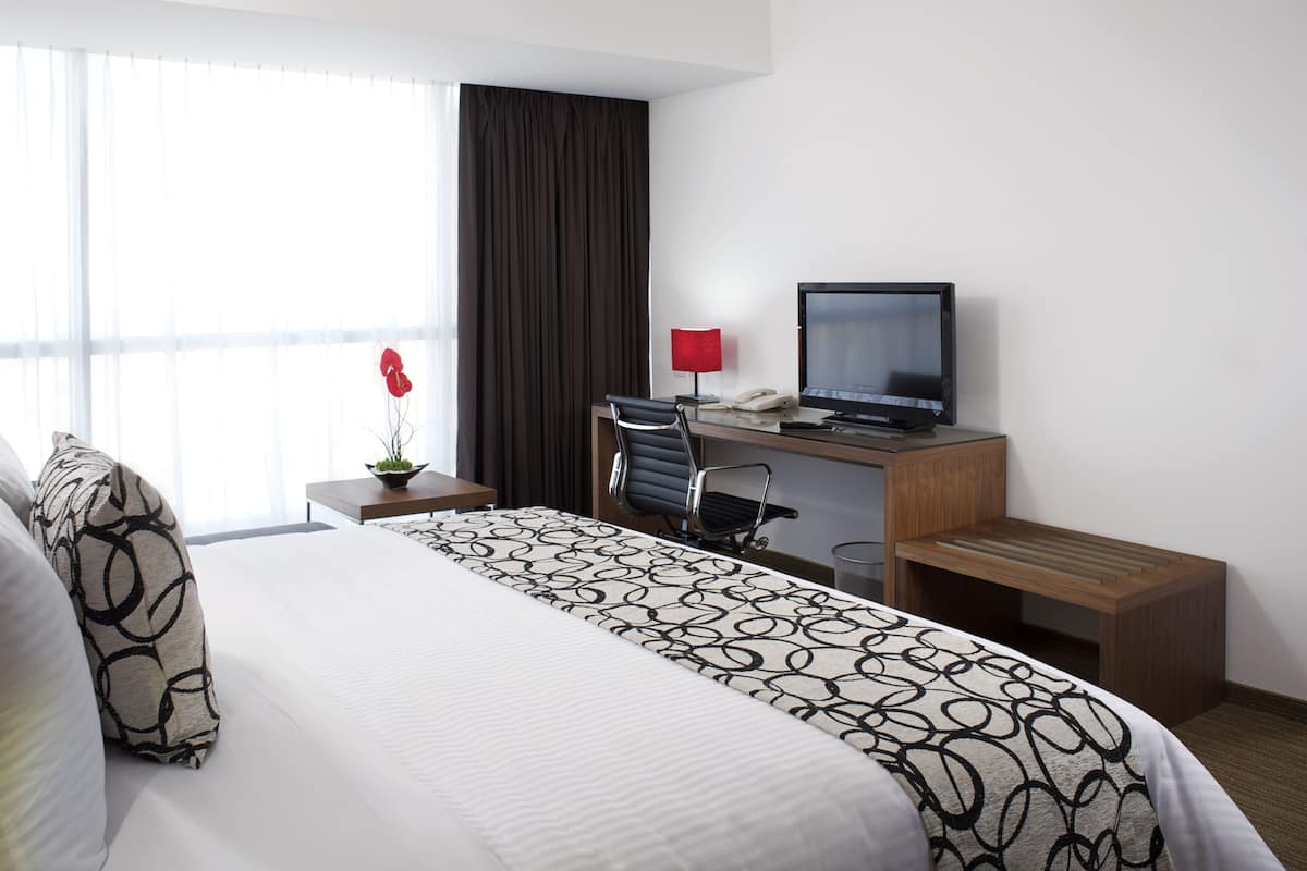 A hotel room with a large bed, a desk with a computer, a chair, a small table with a lamp, and a window with curtains.