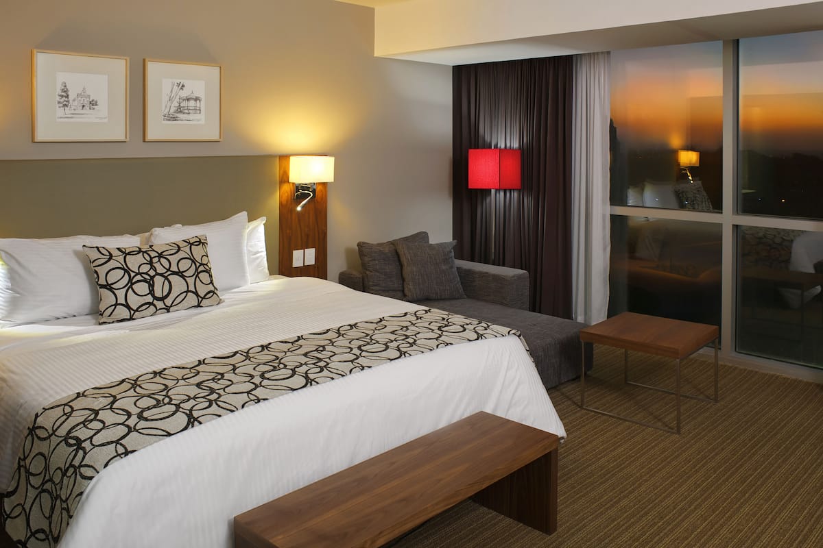 A hotel room with a large bed, a bedside table, a bench, a sofa, a red accent wall, and a large window with curtains.