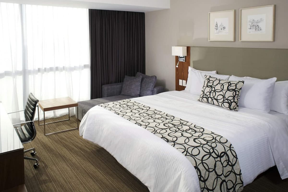 A hotel room with a bed, a desk, a chair, a large window with curtains, and two framed pictures on the wall.
