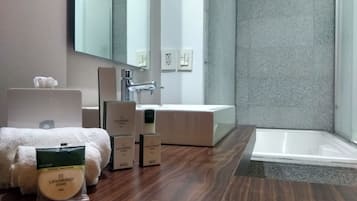 Un baño moderno con suelo de madera, un lavabo blanco, un espejo y una bañera. Hay artículos de aseo en el suelo.