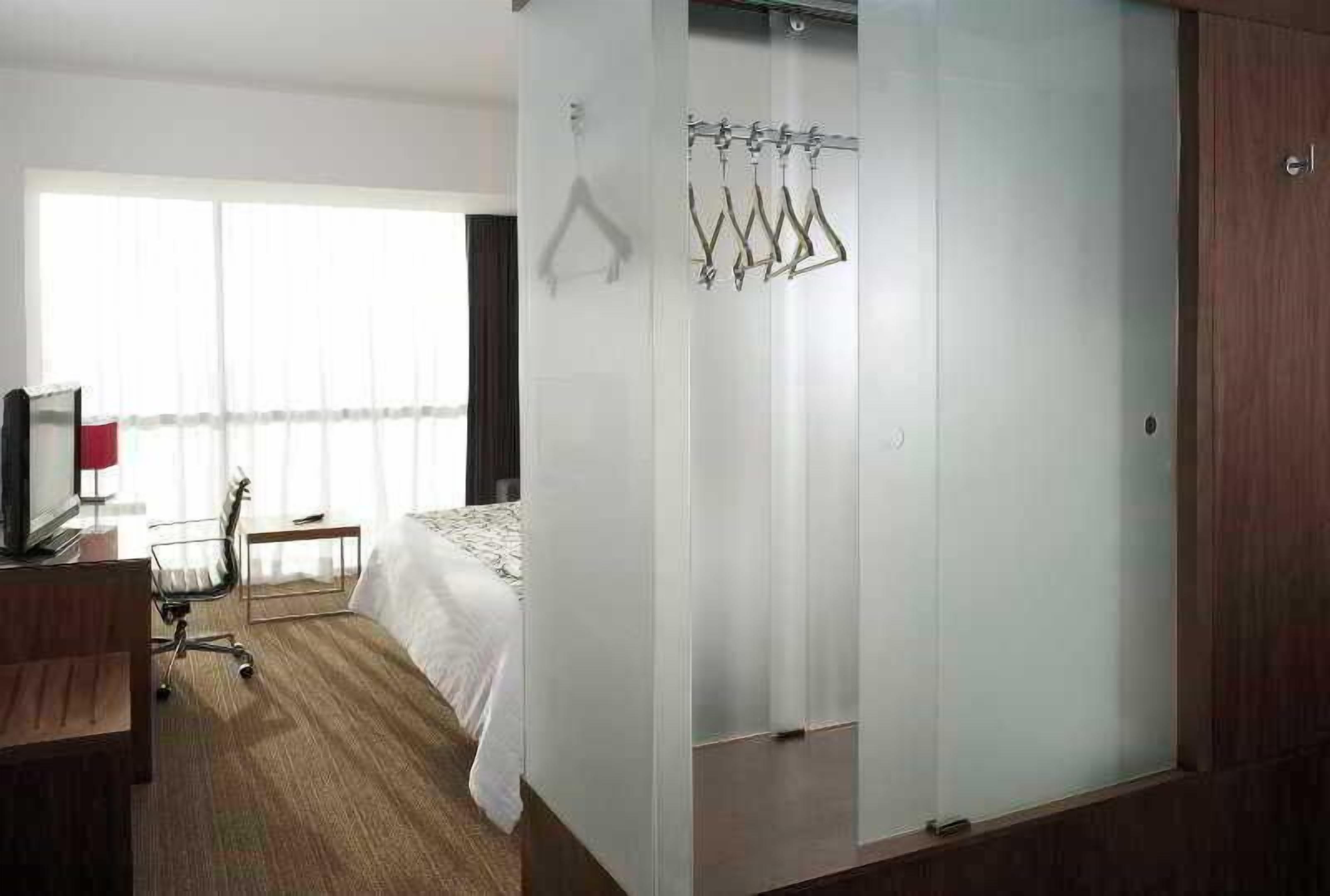 Quarto de hotel com cama, escrivaninha, cadeira, televisão e janela com cortinas.