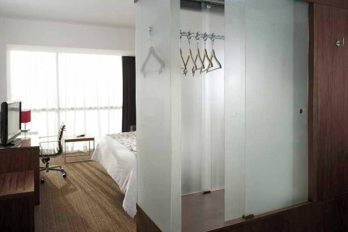 Quarto de hotel com cama, escrivaninha, cadeira, televisão e janela com cortinas.