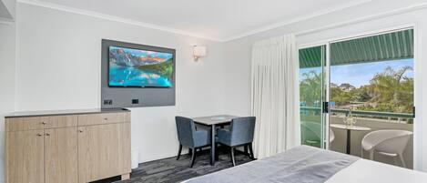 Habitación de hotel con cama, televisión en la pared, una mesita con dos sillas, un armario de madera y un balcón con vistas al exterior.