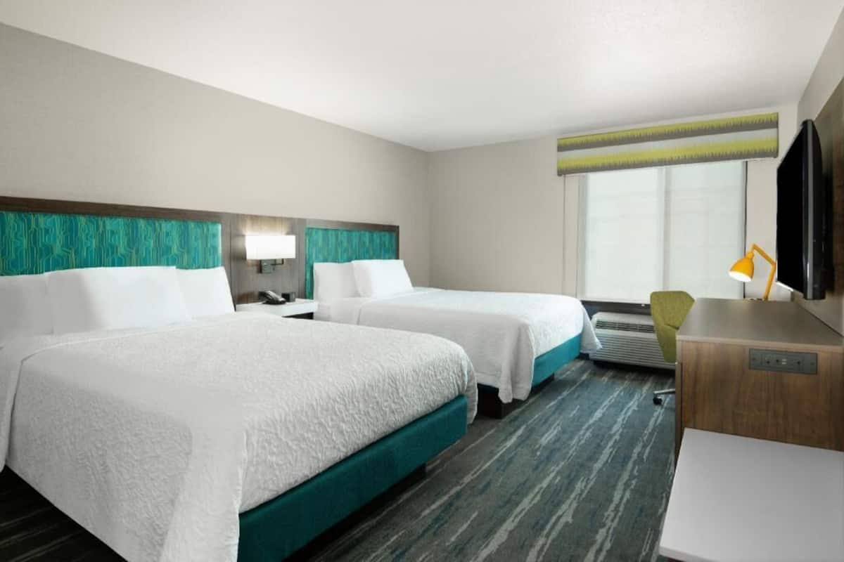 A hotel room with two beds, a desk, a chair, a TV, and a window with blinds.