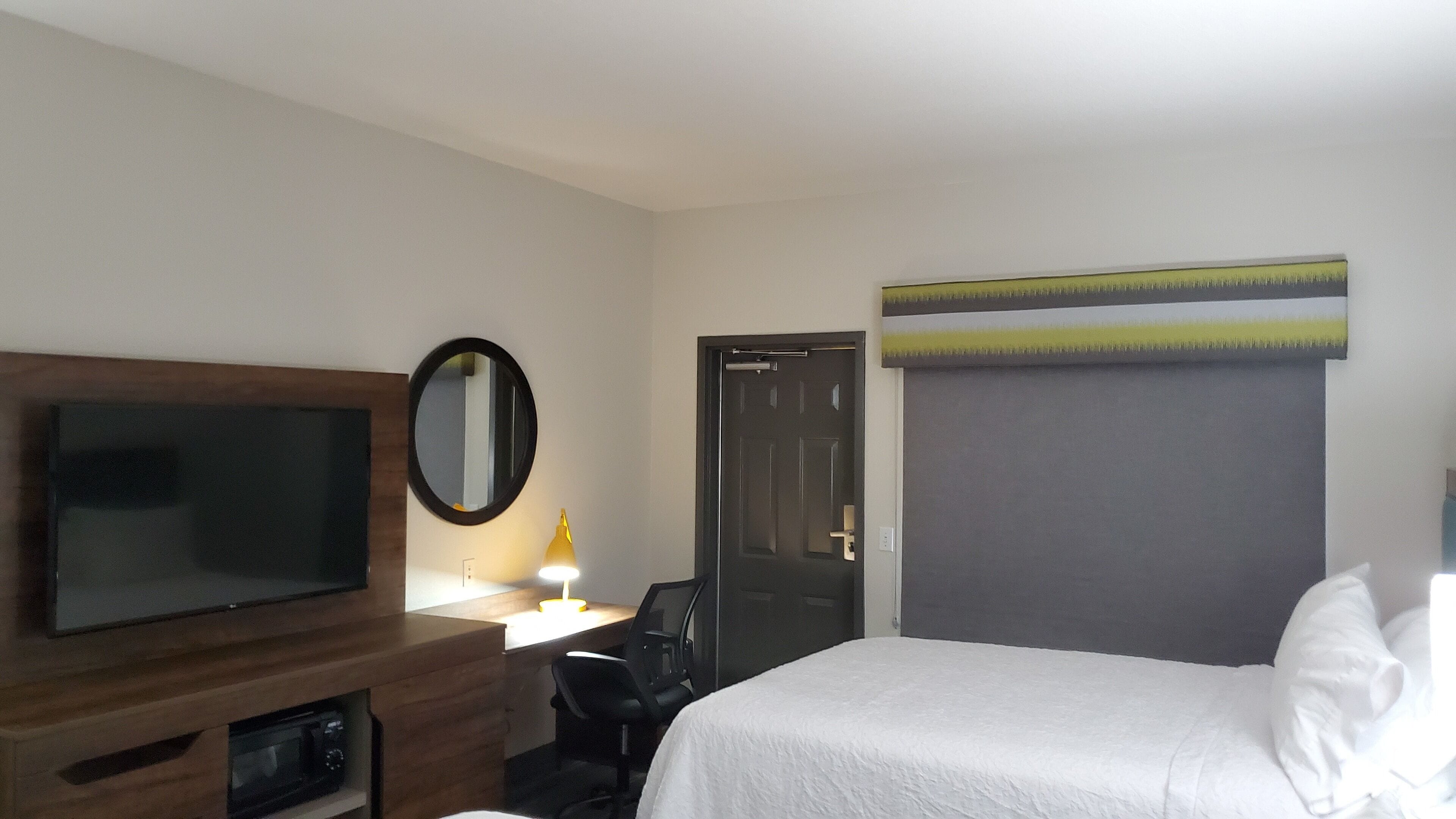 A hotel room with a bed, a TV, a desk with a lamp, and a door.
