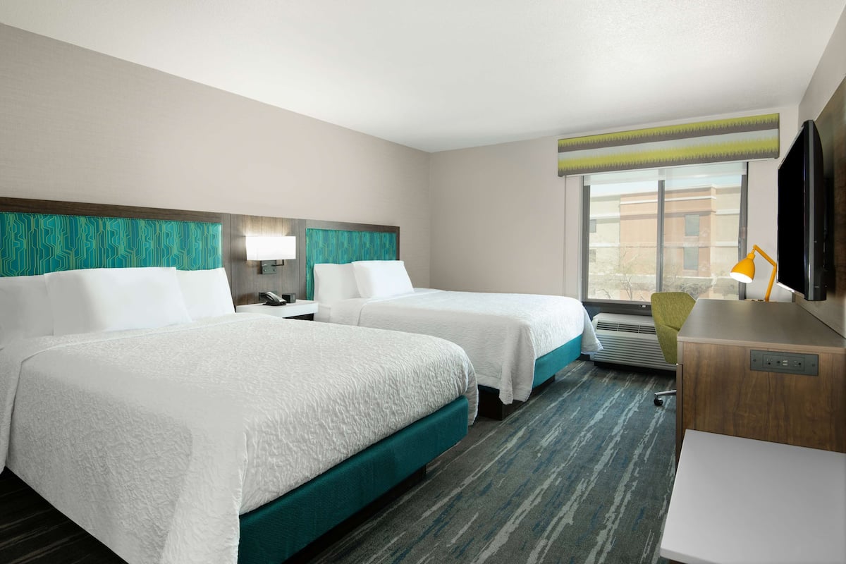 A hotel room with two beds, a desk, a chair, a TV, and a window with a view.