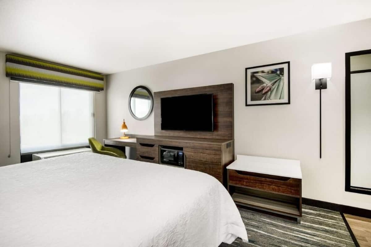 A hotel room with a large bed, a flat-screen TV, a desk with a lamp, a mirror, and a framed picture on the wall.