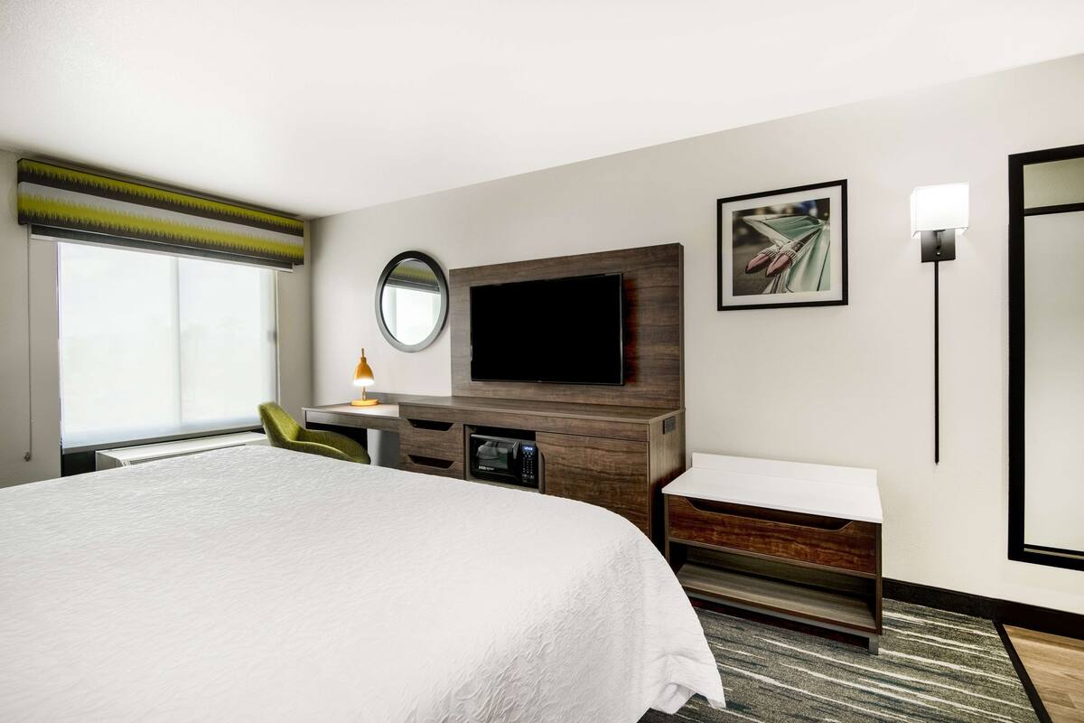 A hotel room with a large bed, a flat-screen TV mounted on a wooden panel, a desk with a lamp, a chair, and a small side table.