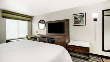 A hotel room with a large bed, a flat-screen TV mounted on a wooden panel, a desk with a lamp, a chair, and a small side table.