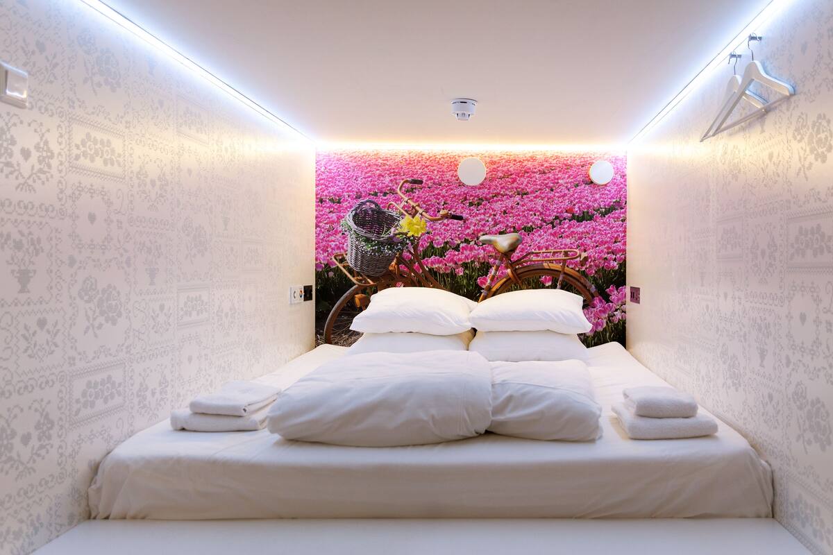 A neatly made bed with white linens and pillows, a wall with a floral wallpaper design, and a bicycle with a basket against the wall.