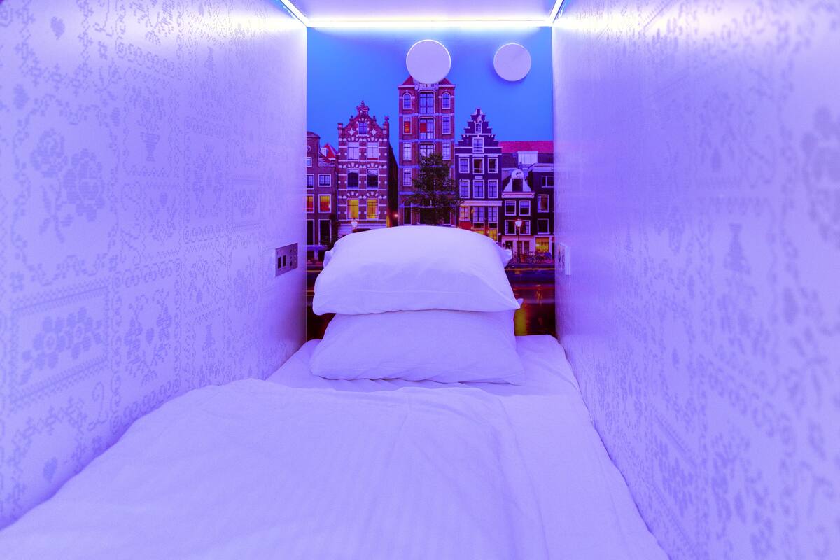 A narrow room with a bed, a wall-mounted light, and a cityscape mural.