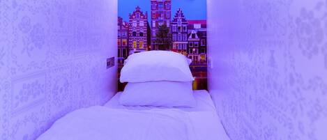 Una habitación estrecha con una cama, una lámpara de pared y un mural de paisaje urbano.