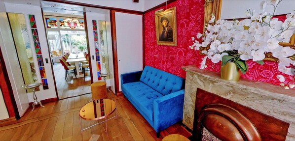 Une pièce avec un canapé bleu, un plancher en bois, un miroir et un mur rouge à motifs, orné d’un portrait encadré.