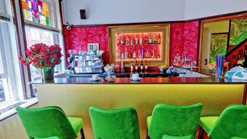 A bar area with a red patterned wall, a coffee machine, and a variety of bottles. There are green chairs in front of the bar.