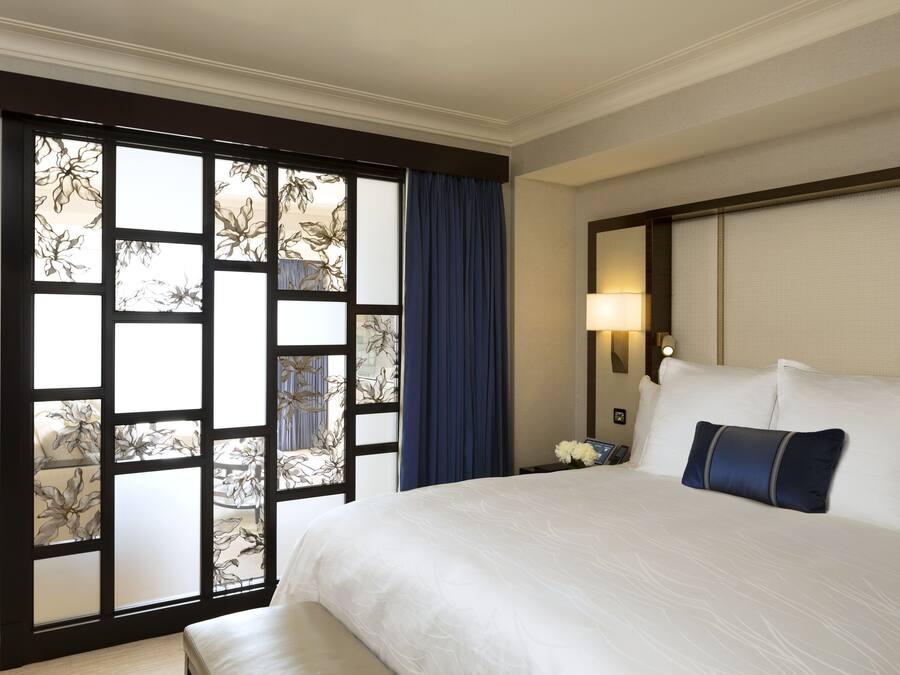 A hotel room with a large bed, a nightstand, a lamp, and a view through a window with a floral pattern.