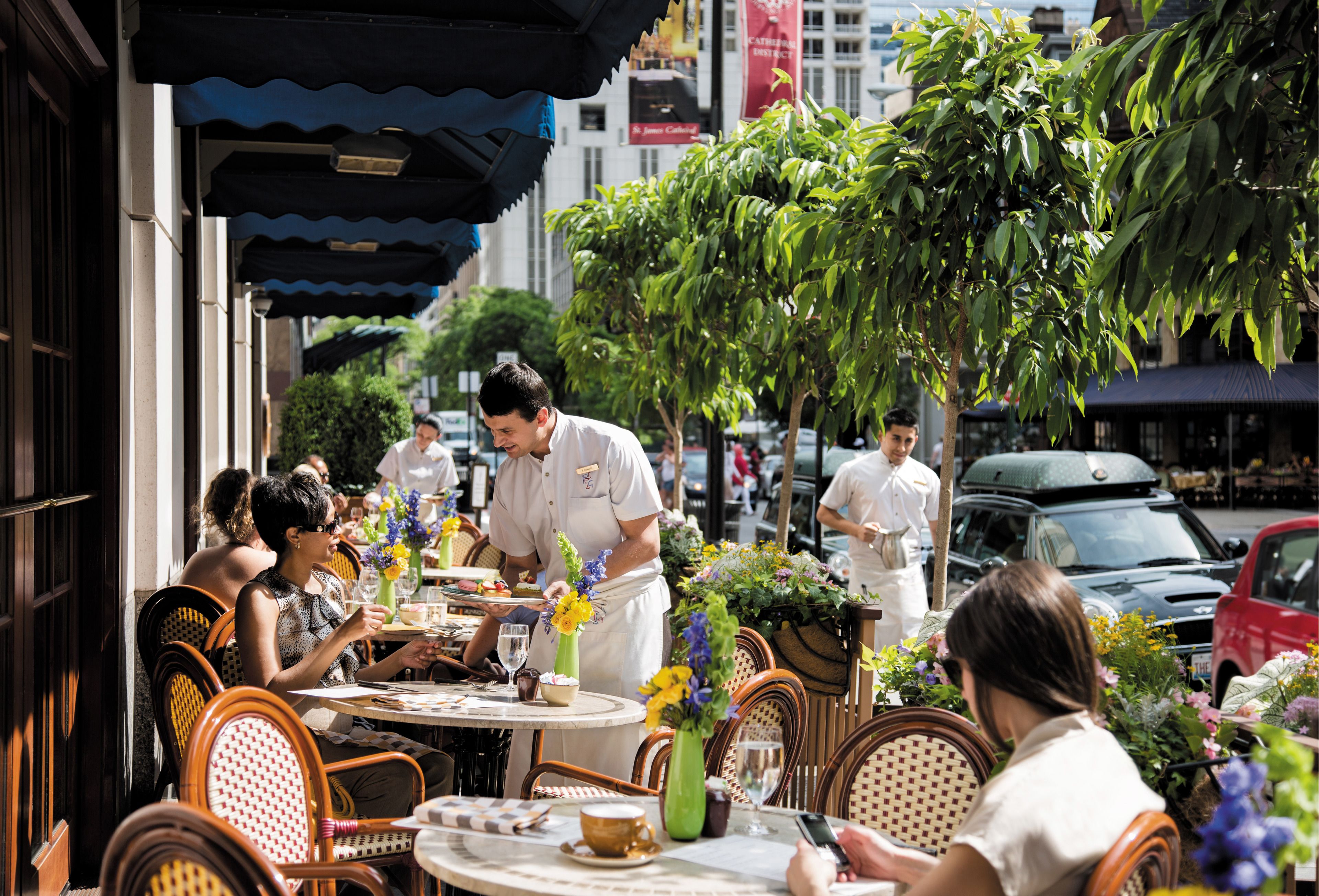 An outdoor café with wicker chairs and tables, a waiter serving food, and a street view with cars and buildings in the background.