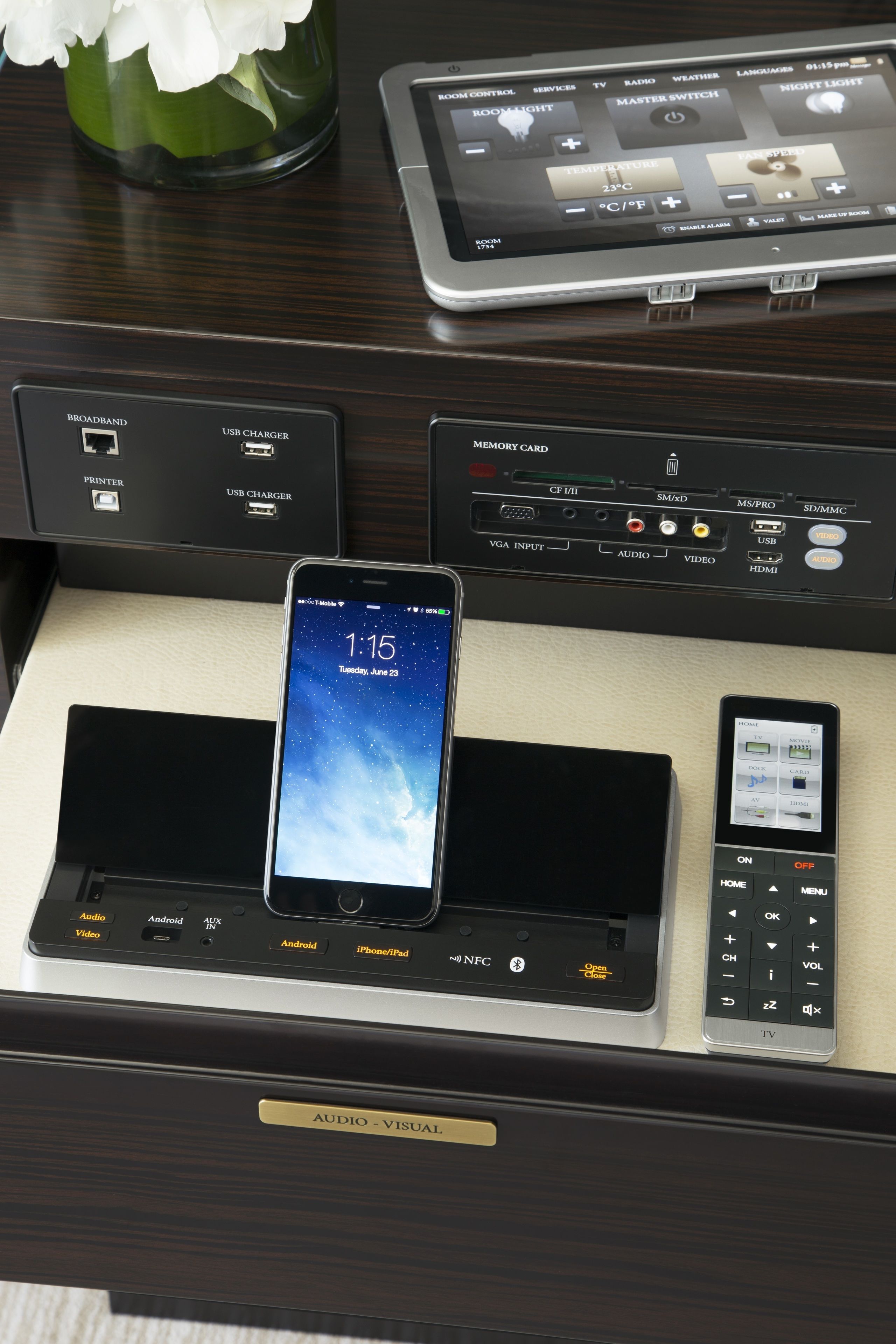 Un panneau de commande dans une chambre d’hôtel, avec un smartphone sur une station de recharge, une télécommande et un téléviseur.