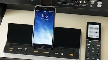 Un panneau de commande dans une chambre d’hôtel, avec un smartphone sur une station de recharge, une télécommande et un téléviseur.
