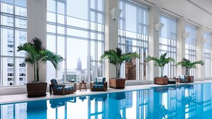 Un área de piscina interior moderna con ventanas amplias, palmeras en macetas y sillones de descanso.