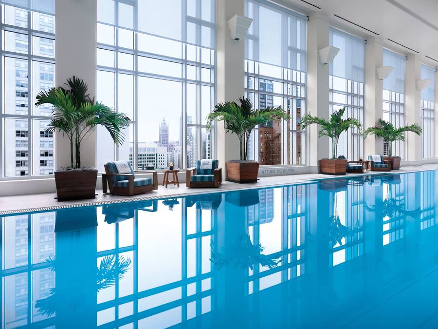 A modern indoor pool area with large windows, potted palm plants, and lounge chairs.