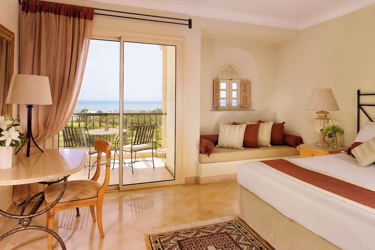A hotel room with a bed, a desk with a chair, a small table, a sofa, and a view of the sea through a sliding door.