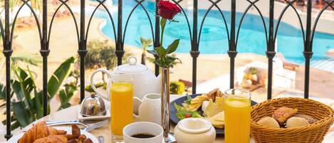 Une table de petit-déjeuner avec des croissants, des œufs et du jus, donnant sur une piscine et des palmiers.