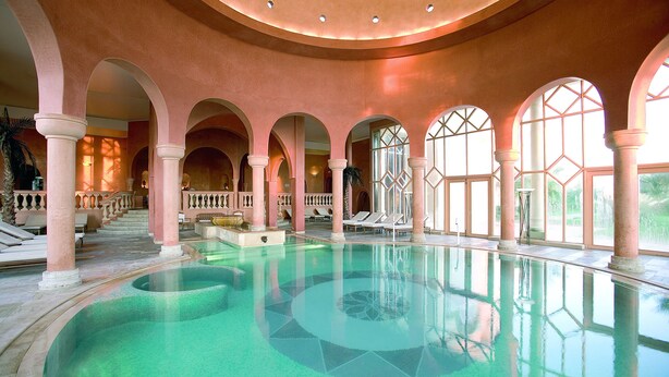 A large indoor swimming pool with clear water, surrounded by arches and columns, and a staircase leading to an upper level. - The Residence Tunis (La Marsa)