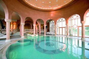 Une vaste piscine intérieure à l’eau claire, entourée d’arcades et de colonnes, et un escalier menant à un niveau supérieur.